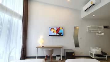 A hotel room with two beds, a desk with a chair, a television, an air conditioning unit, a waste bin, and a storage unit with a hanging rack.