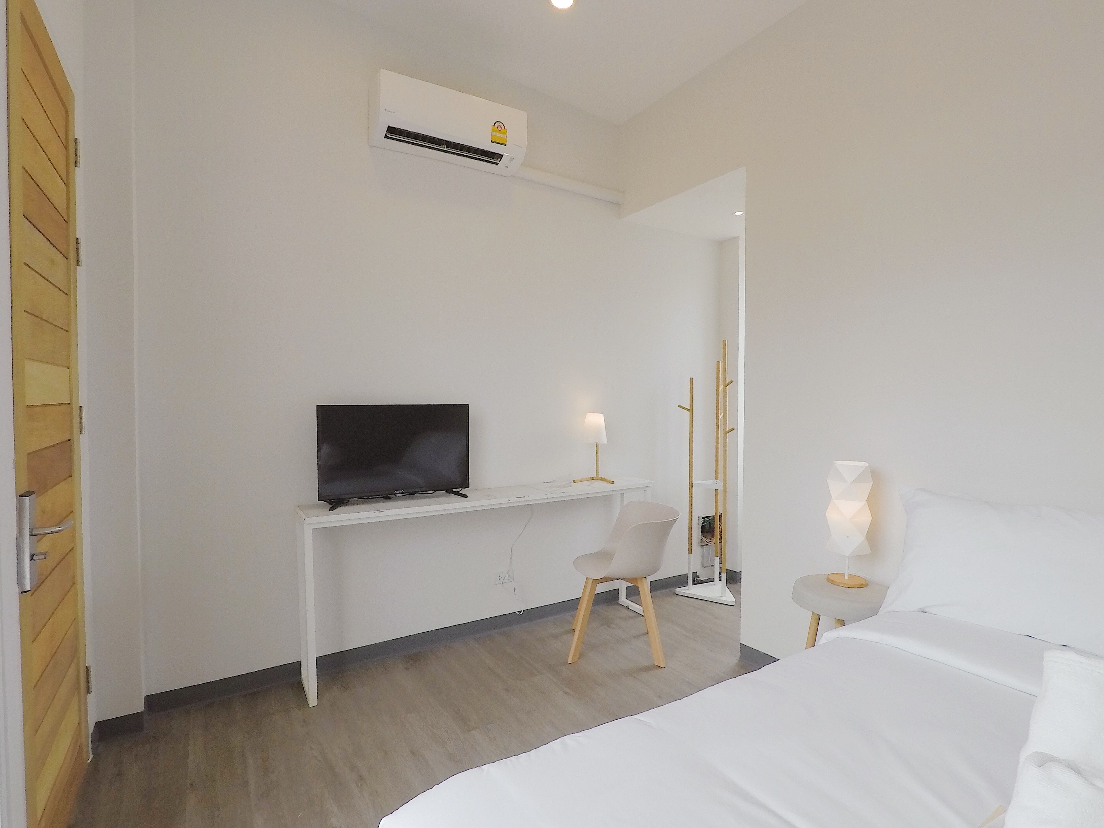 A modern hotel room with a bed, a desk with a flat-screen TV, a chair, a bedside table with a lamp, and an air conditioning unit on the wall.