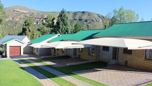 A row of brick buildings with green roofs and white awnings, set against a mountainous backdrop. - La Bri Self Catering (Clarens)