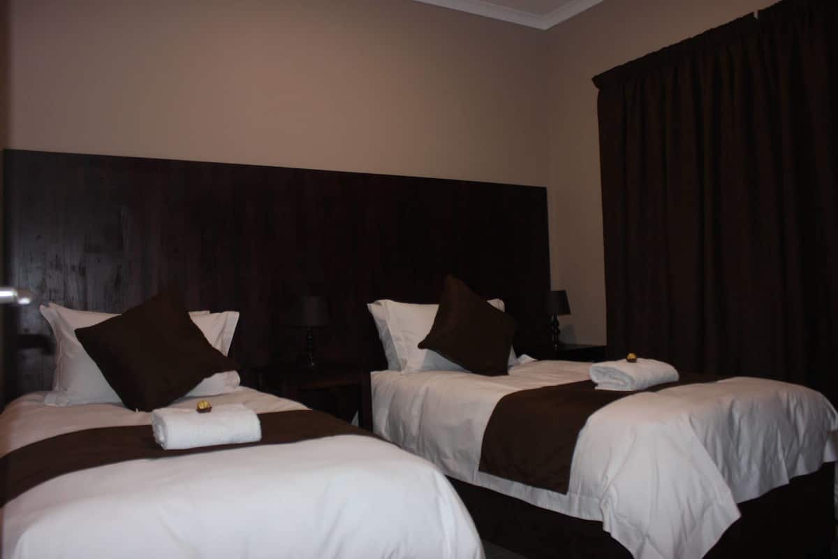 A hotel room with two beds, each with white bedding and brown throw pillows. There is a dark wooden headboard behind the beds, and a large window with dark curtains.