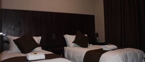 Habitación de hotel con dos camas, cada una con ropa de cama blanca y almohadones marrones. Se observa un cabecero de madera oscura detrás de las camas, y una ventana grande con cortinas oscuras.