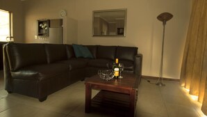 A living room with a dark leather sectional sofa, a wooden coffee table with a bottle and glass, a floor lamp, and a mirror on the wall. - La Bri Self Catering (Clarens)