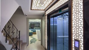 Un pasillo con un ascensor moderno, una escalera con barandillas decorativas y un diseño de techo con motivos.