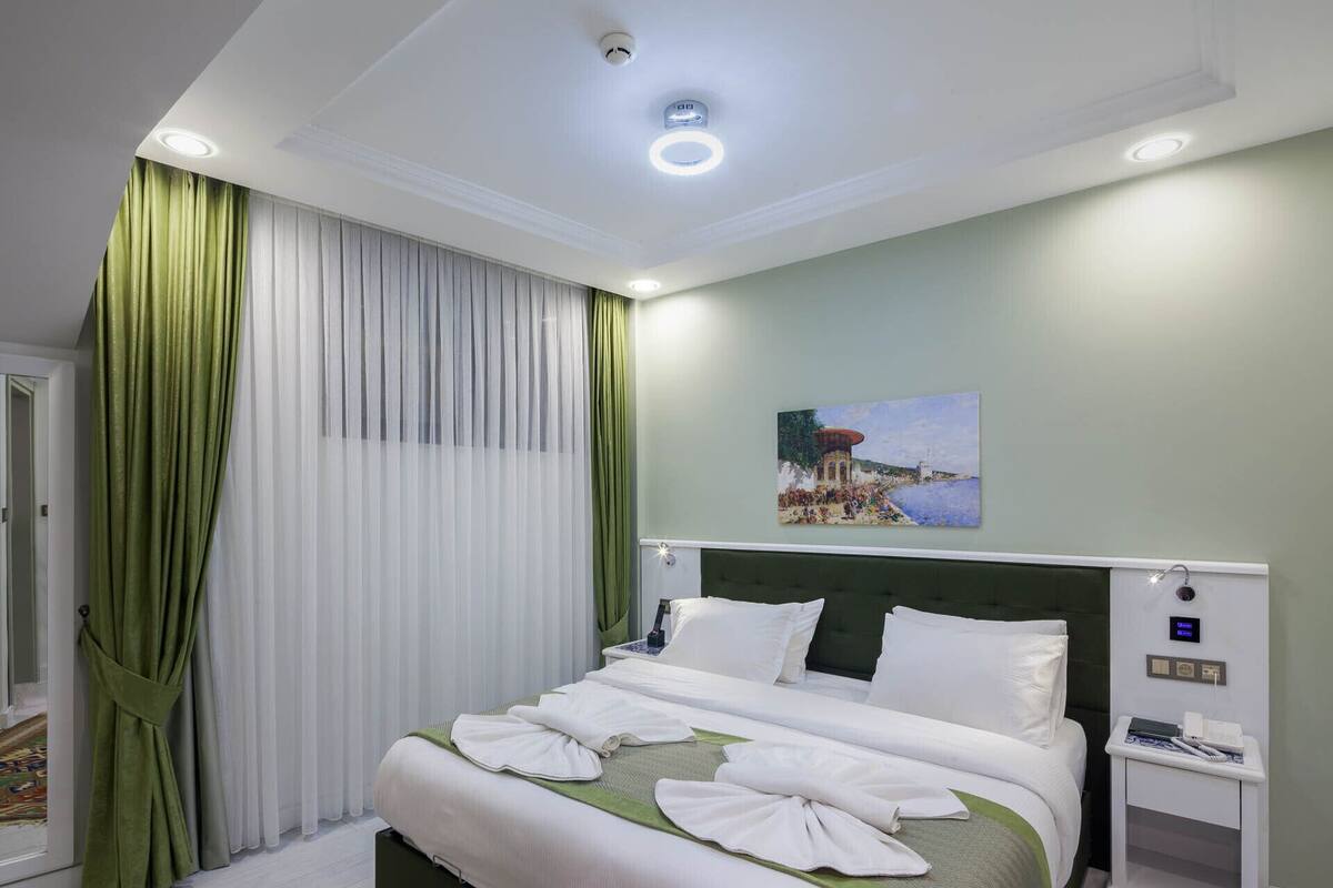 A hotel room with a large bed, green curtains, a painting on the wall, and a small bedside table with a lamp.