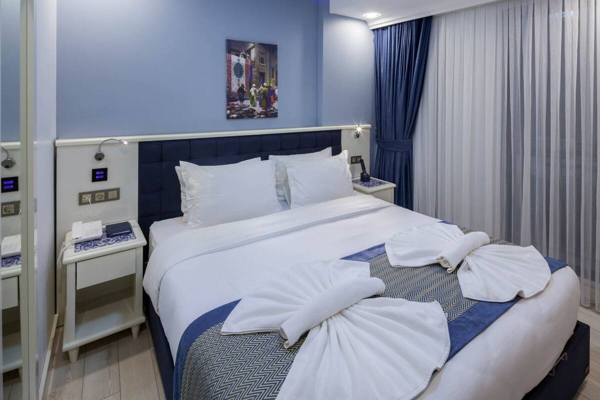 A hotel room with a large bed, a nightstand with a phone and a lamp, a ceiling light, a wall-mounted clock, and a window with curtains.