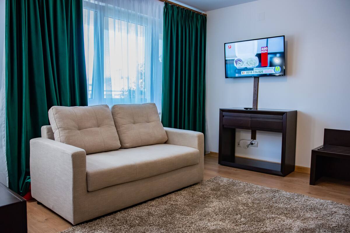 A living room with a beige sofa, a TV on a stand, and a window with green curtains.