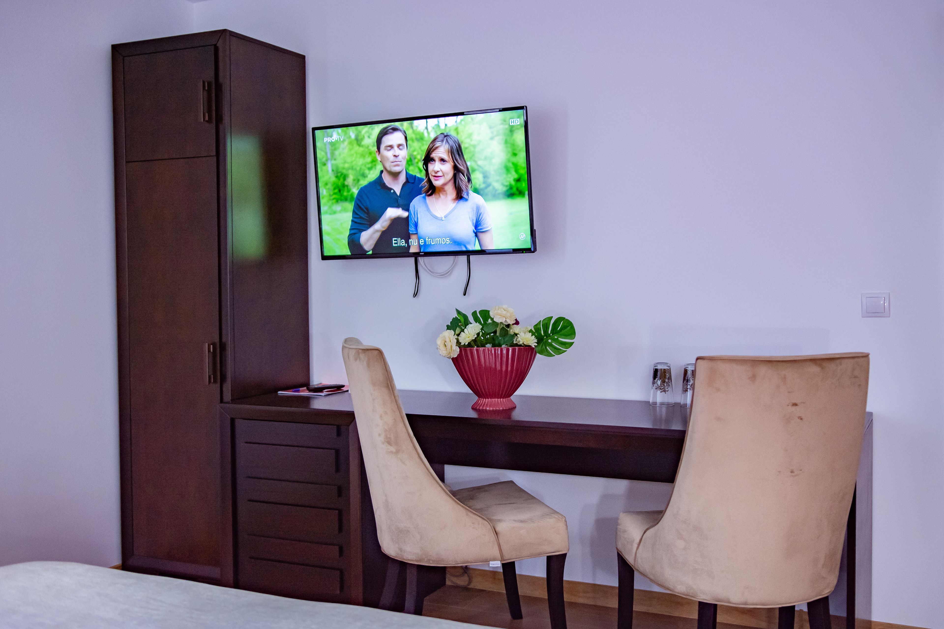 Una habitación con televisión montada en la pared, un escritorio de madera, dos sillas, un jarrón rojo con flores y un armario.