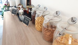 A breakfast bar with various food items and condiments, including cereal and granola, with serving spoons.
