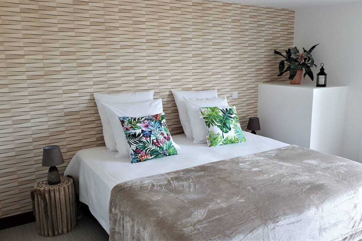 A bedroom with a bed, white pillows, a tropical-themed pillow, a potted plant, a black lantern, and a small wooden stool.
