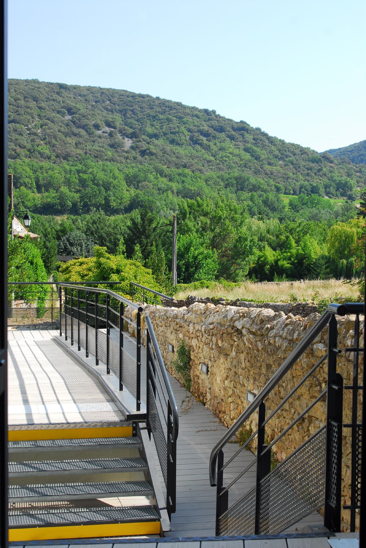 A stone staircase leading up to a bridge with metal railings, surrounded by lush greenery and hills.