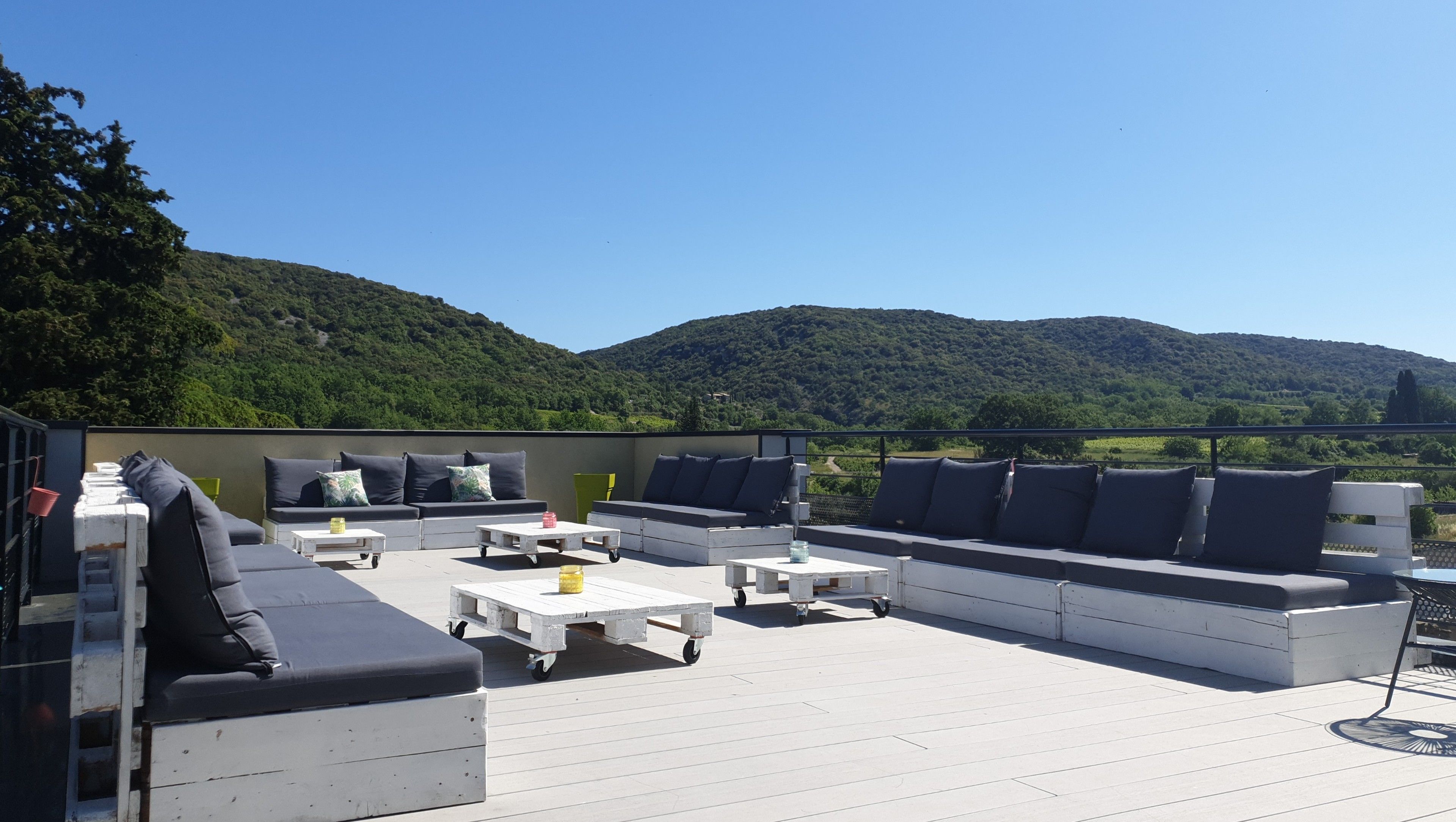 A rooftop terrace with a sofa set, tables, and a view of a green hillside.