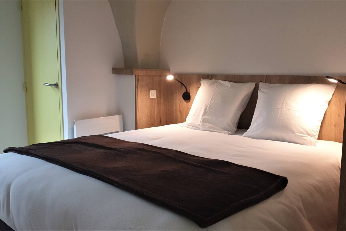 A neatly made bed with white linens and a brown throw blanket, a wooden headboard with wall-mounted lights, and a green door to the left.