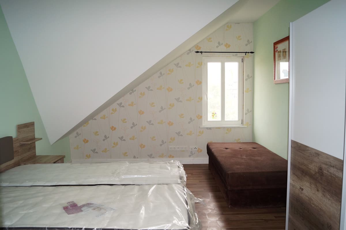 A room with a sloped ceiling, a bed with a mattress cover, a bench, a window with a curtain, and a mirror on the wall.