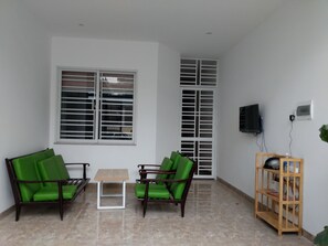 A living room with two green cushioned chairs, a small table, a TV mounted on the wall, and a wooden shelf with electronic devices.