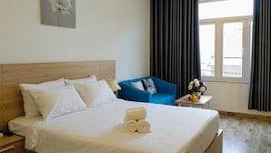 A hotel room with a large bed, a blue armchair, a small table with a vase of flowers, and a window with curtains. - La Maison De Ruby Apartment (Da Nang)