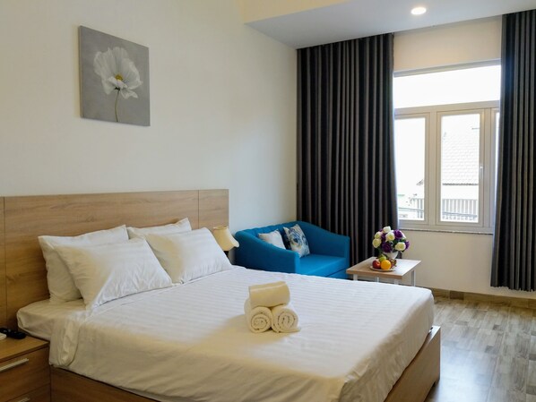 A hotel room with a large bed, a blue armchair, a small table with a vase of flowers, and a window with curtains.