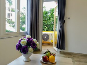 Um quarto com janela, um vaso branco com flores roxas e brancas, e um prato com frutas.
