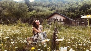 A person sitting in a field of daisies with a wooden cabin in the background. - Farma Sotira (Leskovik)
