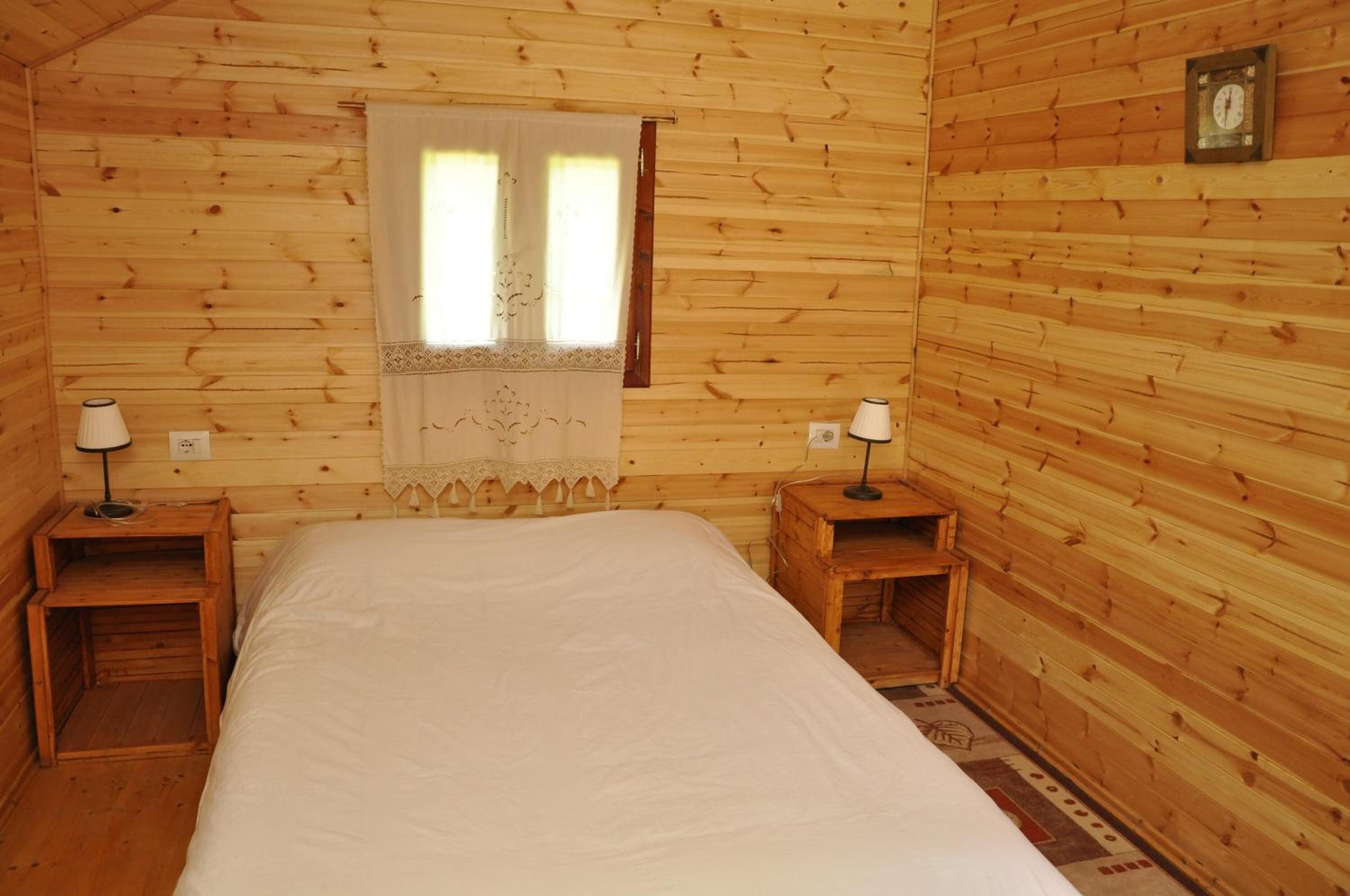 Une chambre rustique en bois, avec un lit, deux tables de chevet, une fenêtre avec des rideaux et une horloge au mur.
