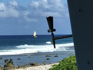 Blick auf das Meer mit einem Segelboot in der Ferne, felsiger Küstenstreifen und ein Gebäude mit schrägem Dach.