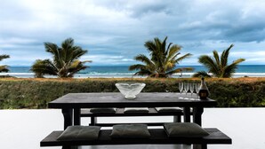 A wooden table with a glass bowl and wine glasses, overlooking a beach with palm trees and the ocean under a cloudy sky. - Coast Cook Islands - 3-Bedroom Beachfront Pool Villa (Rarotonga)