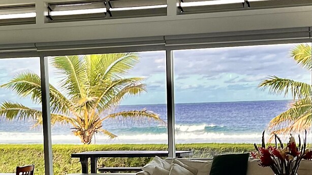 A modern living room with a large window overlooking a beach and ocean. - Coast Cook Islands - 3-Bedroom Beachfront Pool Villa (Rarotonga)