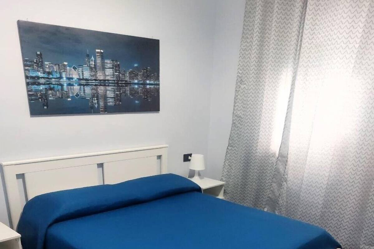 A bedroom with a bed, a nightstand, a lamp, a painting on the wall, and a vase with blue flowers.