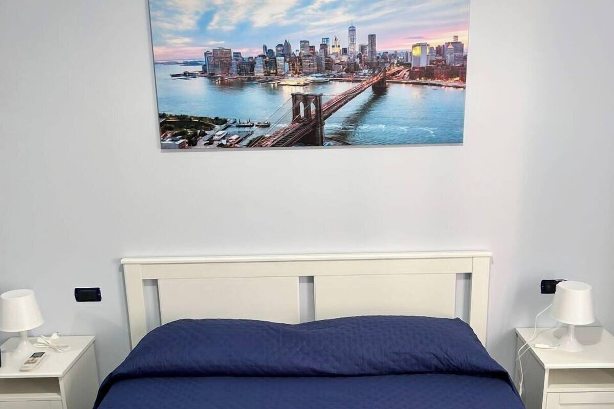 A hotel room with a large bed, white nightstands, a cityscape painting, and two rolled towels on the bed.