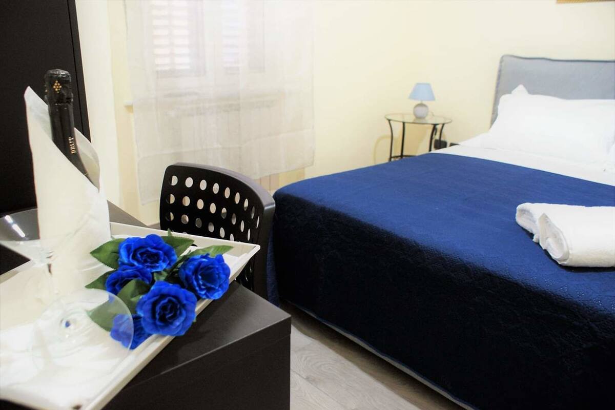 A hotel room with a bed, a desk with a vase of blue roses, a chair, a lamp, and a window with blinds.