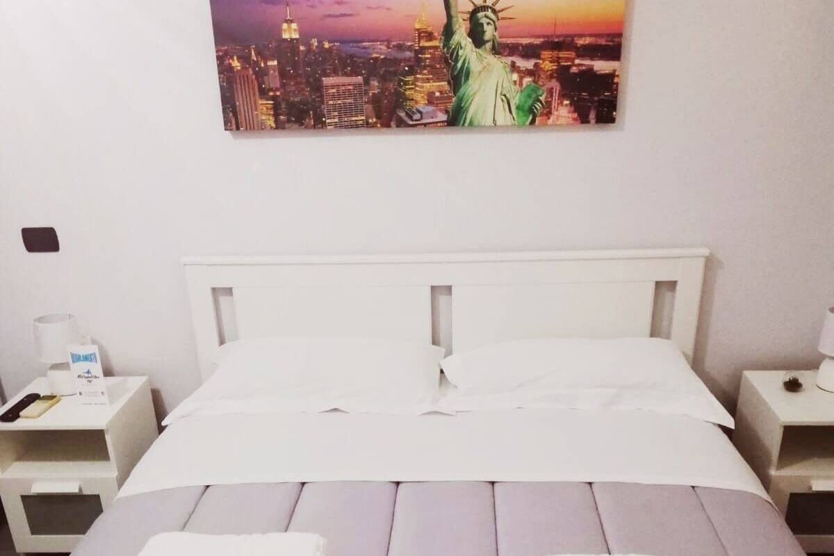 A hotel room with a bed, two white nightstands, and a painting of the Statue of Liberty.