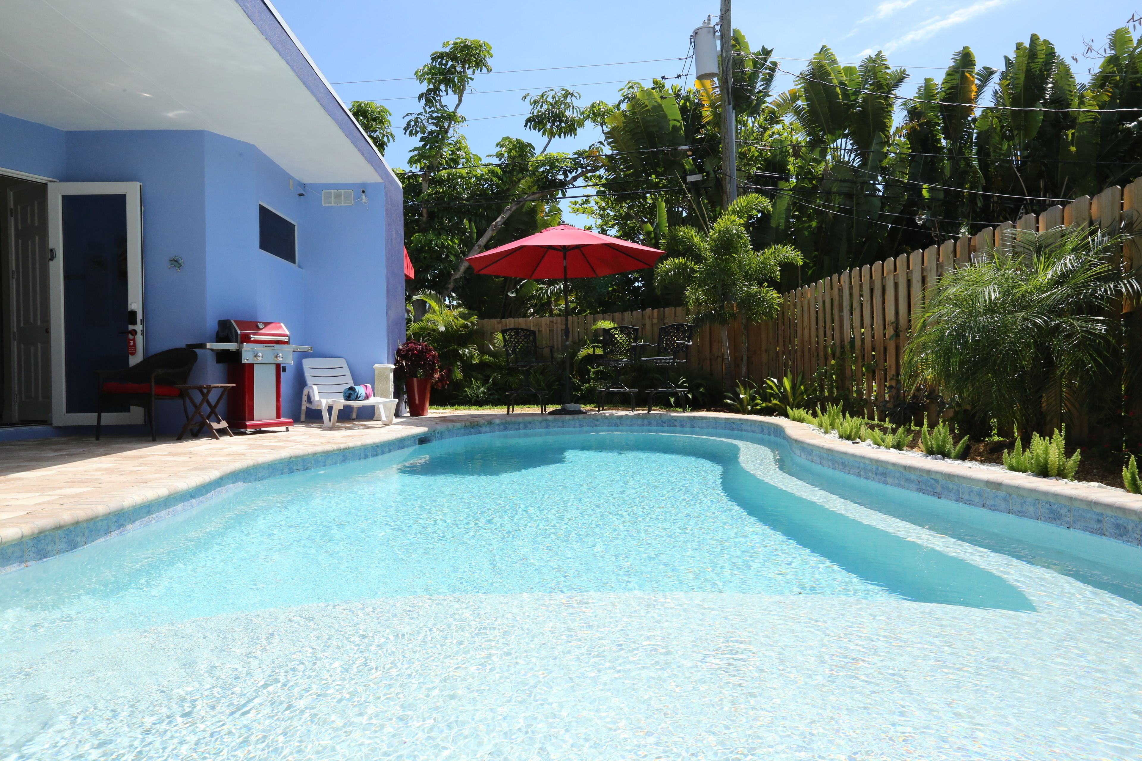 Une piscine avec une terrasse en bois, un barbecue et un parasol rouge.
