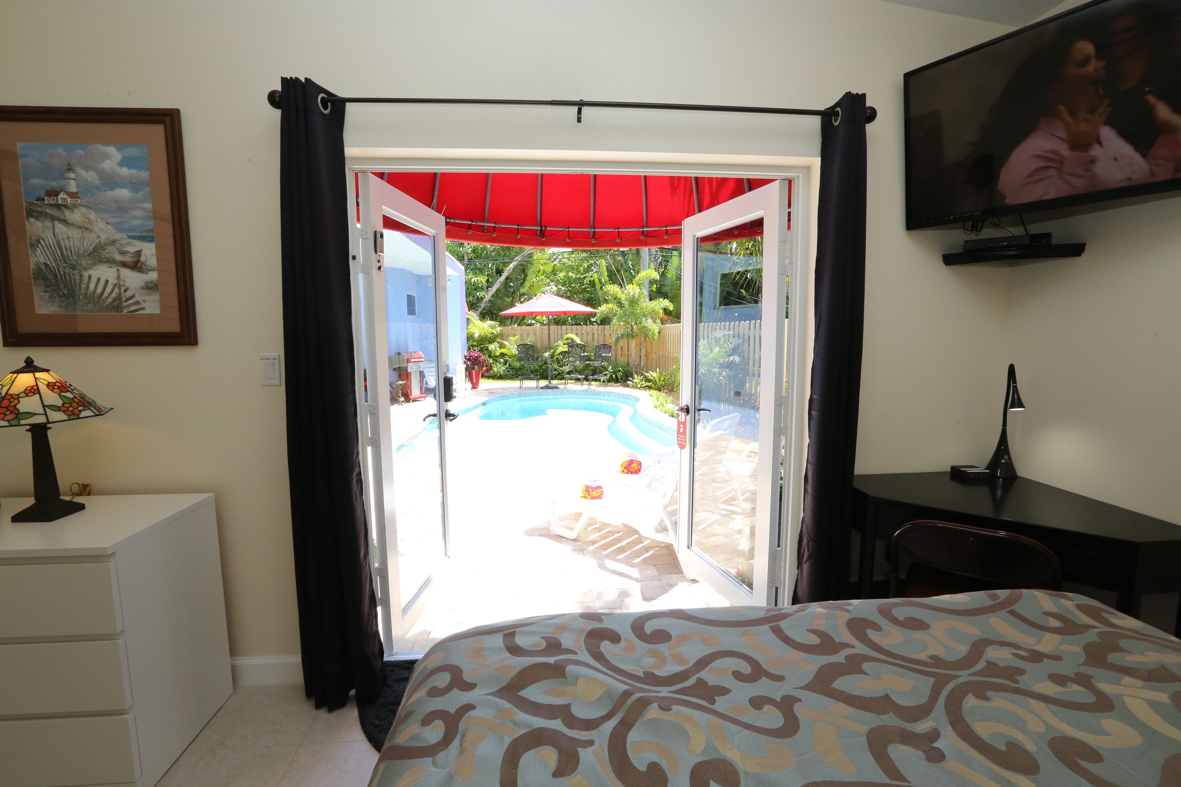 A bedroom with a bed, a desk, a TV, a lamp, and a view of an outdoor swimming pool.
