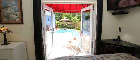 A bedroom with a bed, a desk, a TV, a lamp, and a view of an outdoor swimming pool.