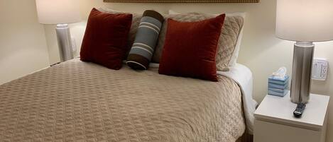 A hotel room with a large bed, two bedside tables, a nightstand, a painting of a beach scene, and a lamp on each table.