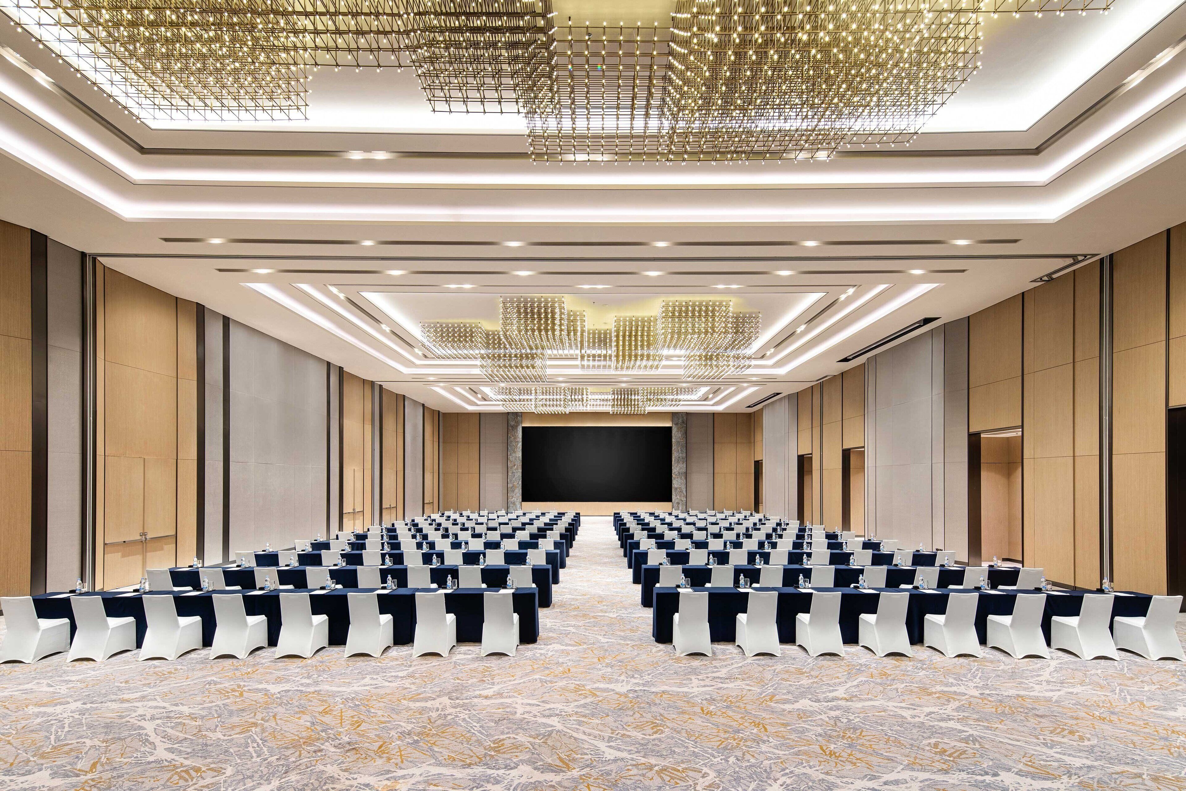 A conference room with rows of tables and chairs, a large screen at the front, and a modern chandelier.