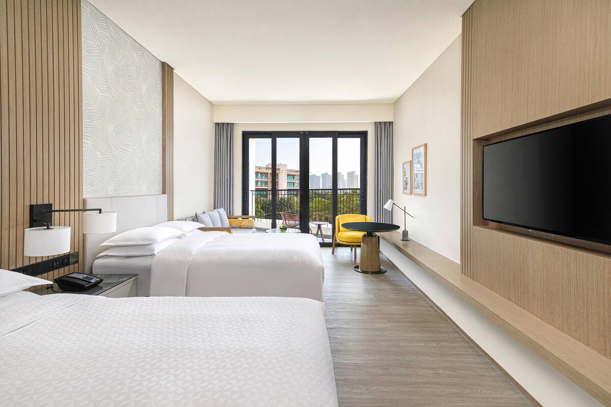 A hotel room with two beds, a TV, a desk with a lamp, and a view of the city.