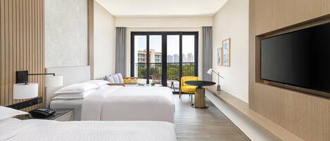 A hotel room with two beds, a TV, a desk with a lamp, and a view of the city.