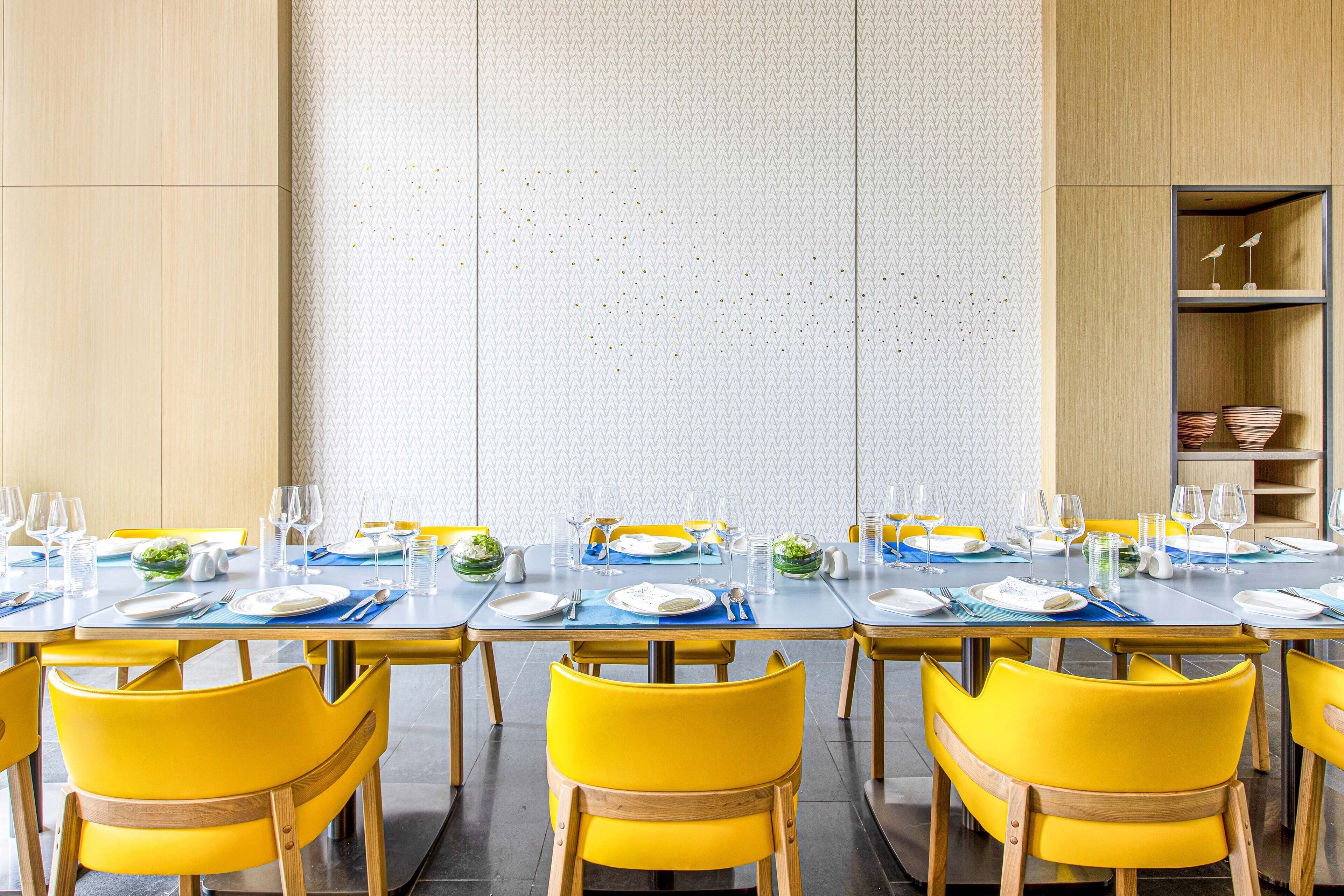 A dining area with a long table set for multiple people, yellow chairs, and a patterned wall in the background.