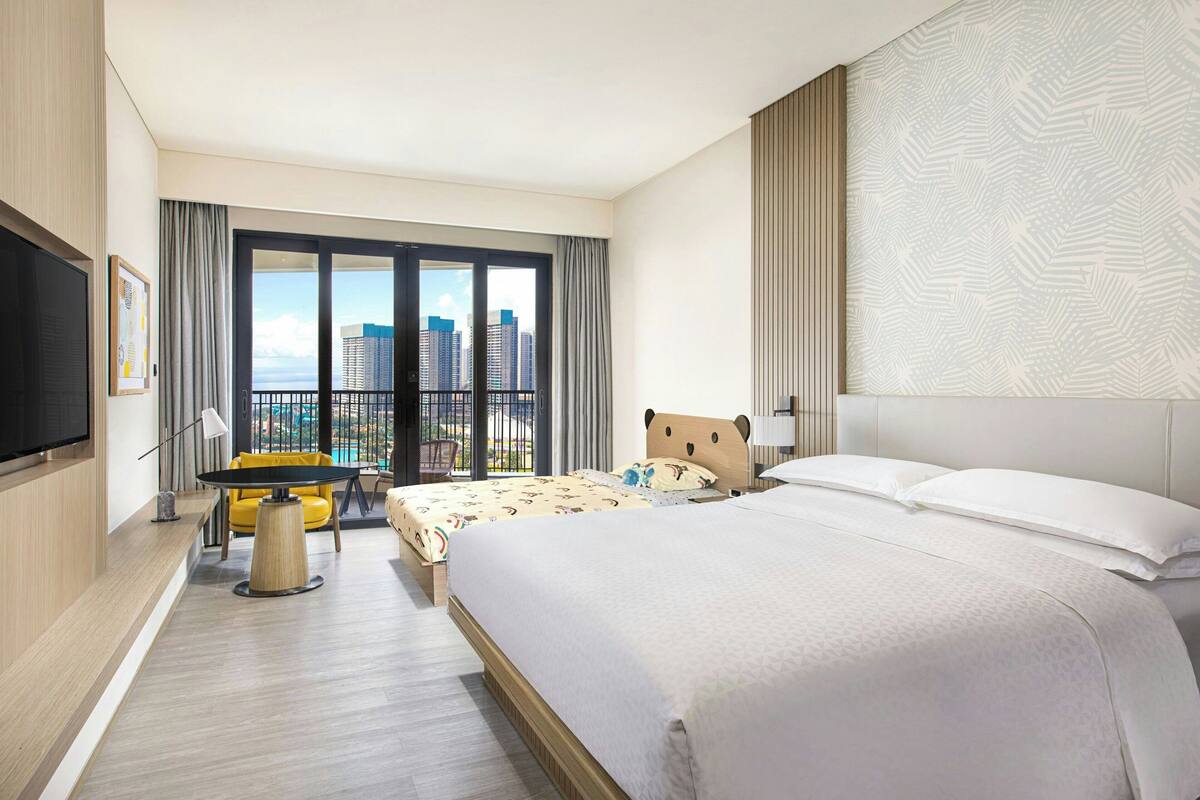 A hotel room with a large bed, a small table, a chair, a TV, and a balcony with a city view.