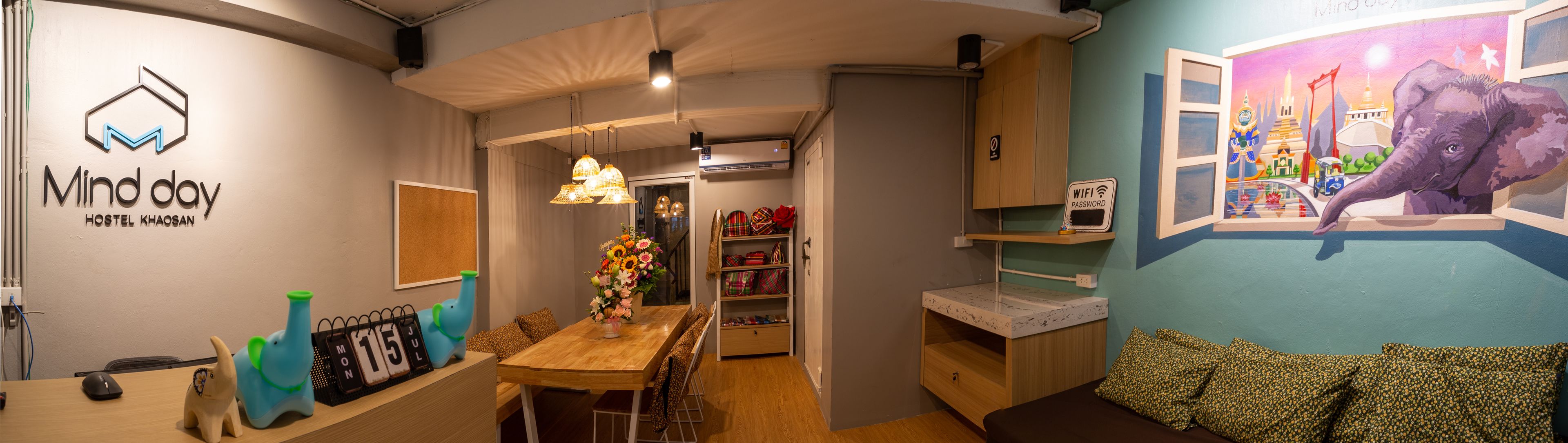 Habitación de albergue con mesa de madera, un calendario, estante en la pared con mochilas y un mural de un elefante.