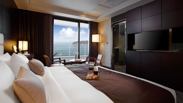 A hotel room with a large bed, a TV, a balcony with a view of the sea, and a dining area with a bottle of wine. - Stellar Hotel (Incheon)