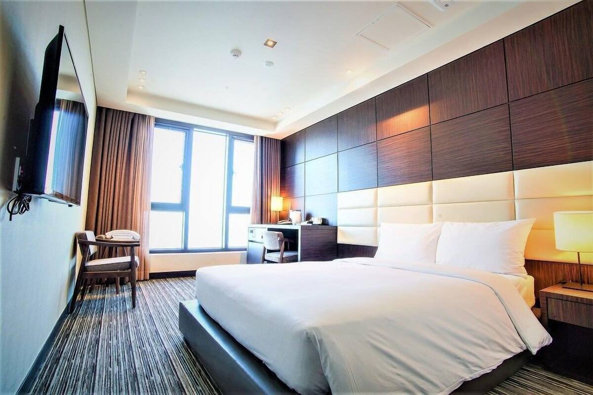 Habitación de hotel con una cama grande, un escritorio, una silla, un televisor y una ventana con cortinas.