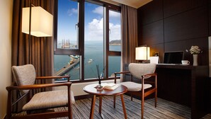 A hotel room with two chairs, a small table, a desk with a laptop, a TV, and a view of a harbor with boats. - Stellar Hotel (Incheon)
