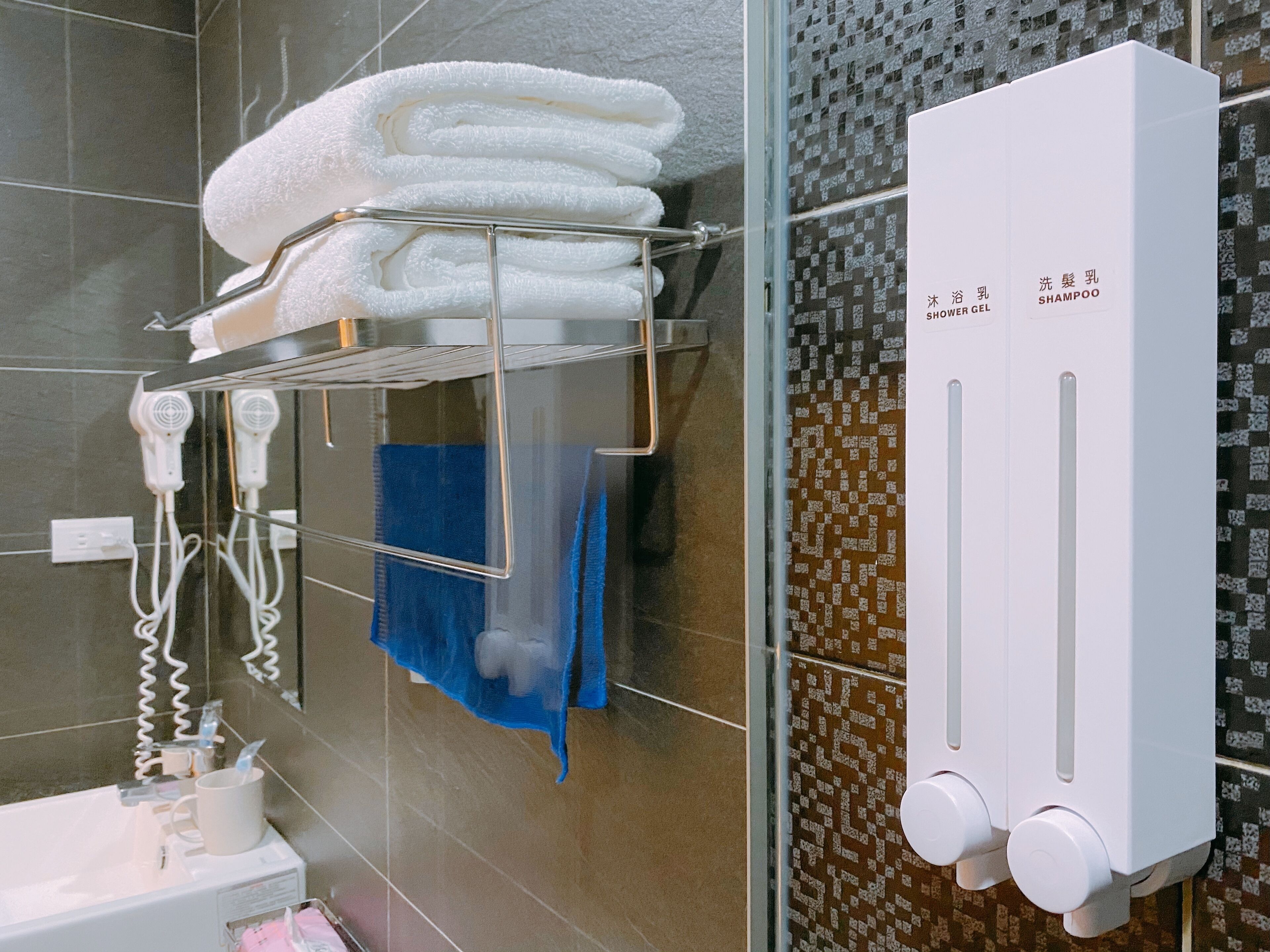 Une salle de bain avec une douche à parois de verre, un porte-serviettes avec des serviettes pliées, une serviette bleue, un sèche-cheveux blanc et un distributeur de gel douche blanc.