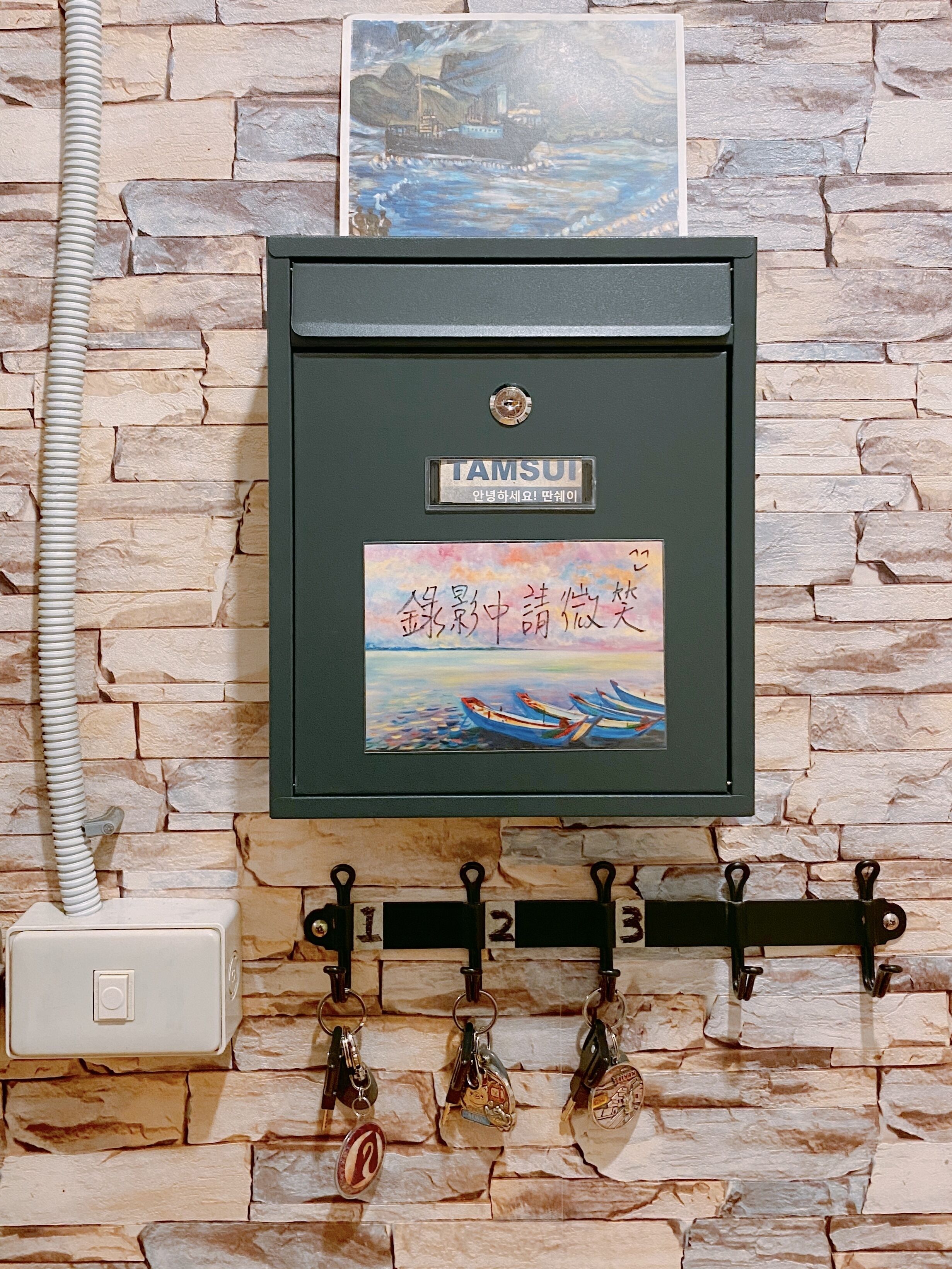 A wall-mounted metal box with a picture of a ship and a painting of boats on water. Below, there are metal hooks with keys and a white electrical outlet.