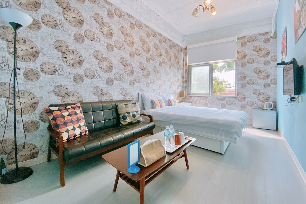 A room with a patterned wallpaper, a black leather sofa, a bed, a small table with bottles, and a floor lamp.