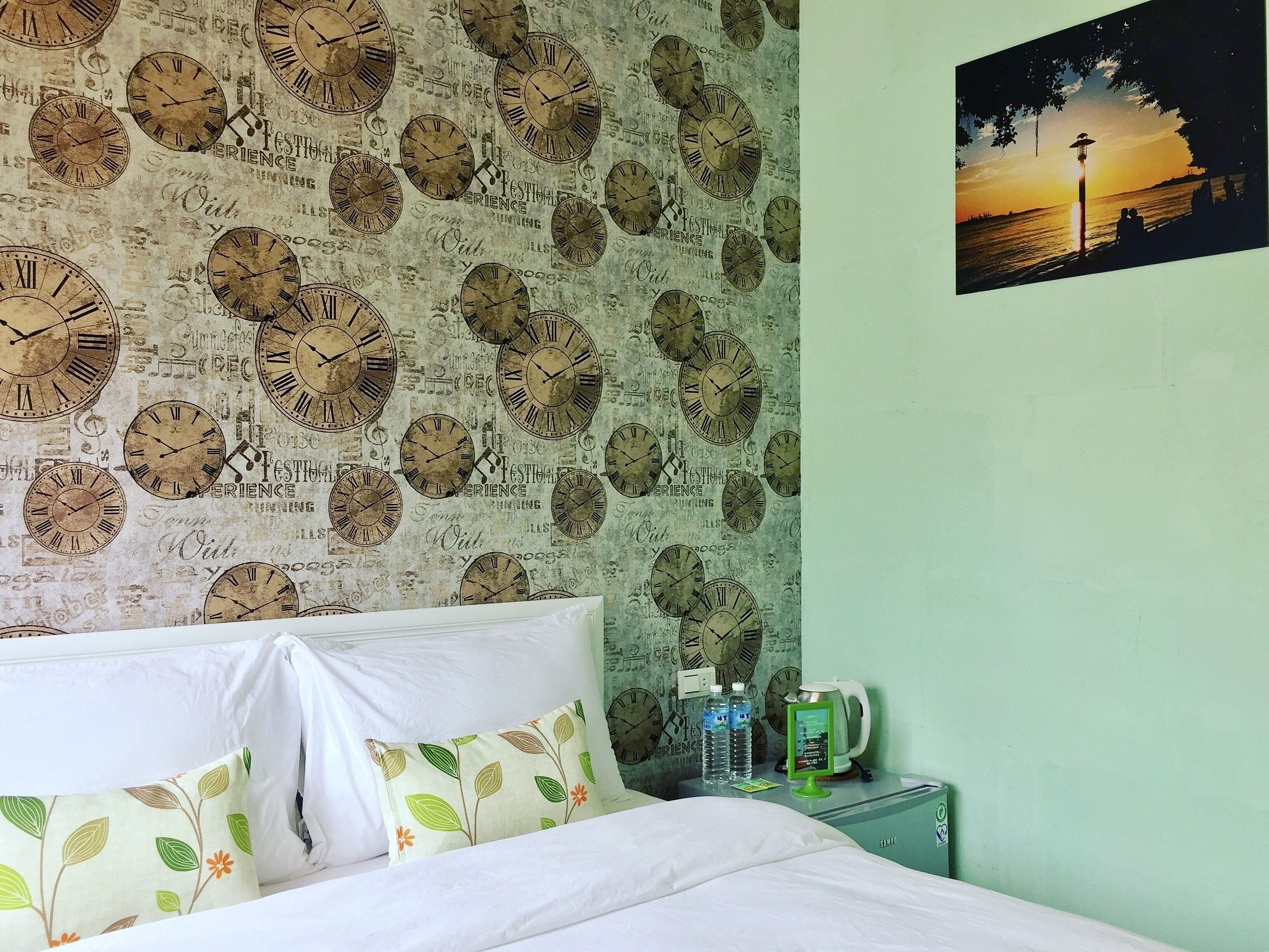 Un dormitorio con una pared llena de relojes, una cama con ropa de cama blanca, una mesita de noche con hervidor y una fotografía de un atardecer.