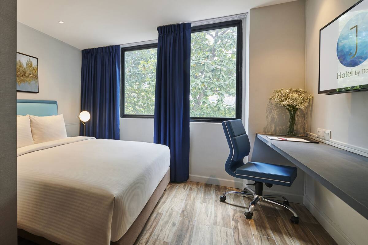 Ein Hotelzimmer mit einem Bett, einem Schreibtisch, einem Stuhl, einer Schreibtischlampe, einer Vase mit Blumen, einem gerahmten Bild an der Wand und einem Fenster mit Vorhängen.