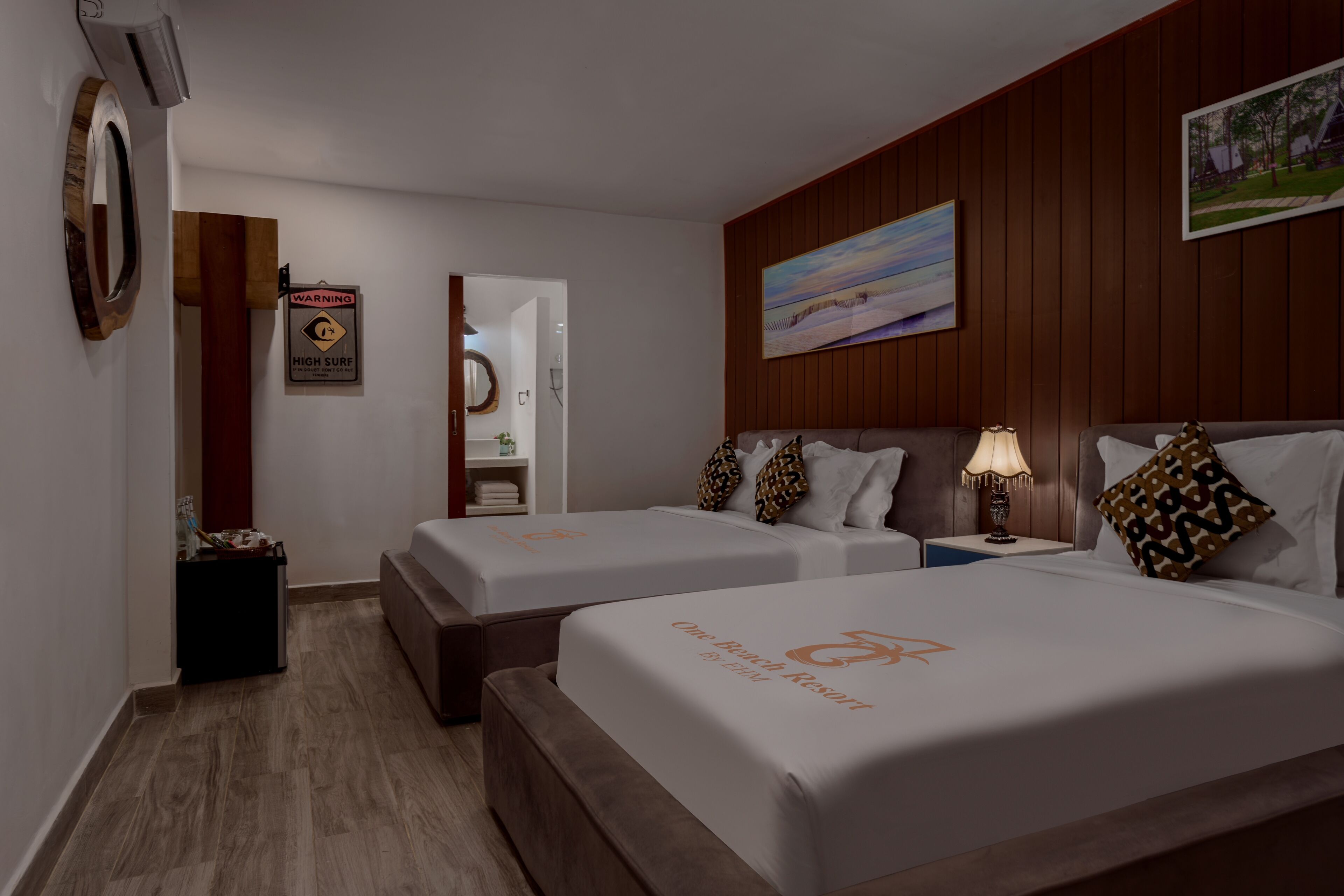 A hotel room with two beds, a wooden panel wall, a painting, a warning sign, a mirror, and a bedside table with a lamp.