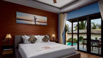 Habitación de hotel con cama, un cuadro, ventilador de techo, mesita de noche, lámpara y vista a un área de piscina al aire libre.